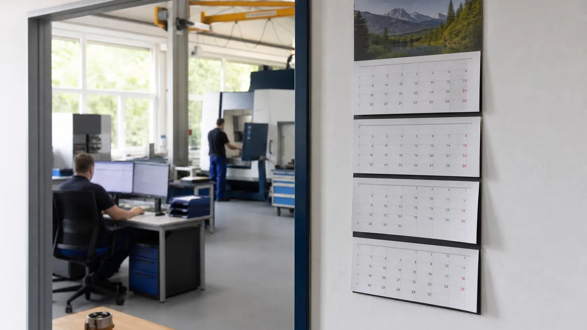 Wandkalender im Werksbüro mit mehreren Monatsblättern, dahinter ein Arbeitsplatz in der Halle